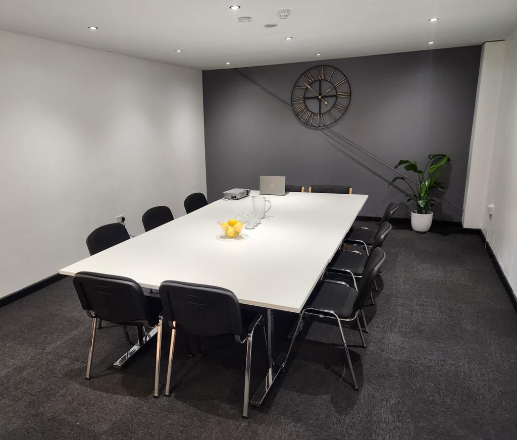 A boardroom with a white conference table, black chairs, wall clock, and potted plant at NO 36.