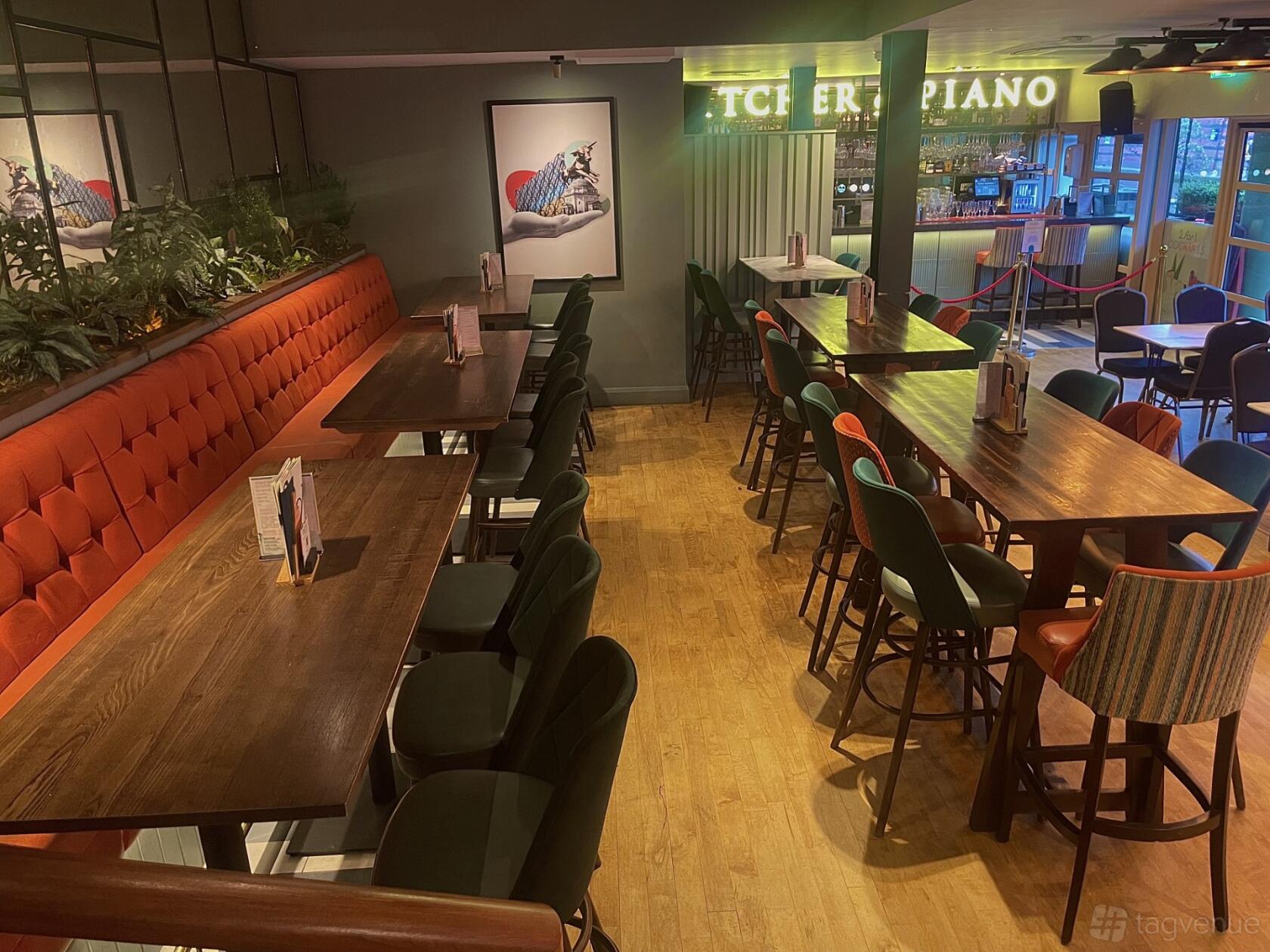 An event space in a bar with long wooden tables, upholstered bench seating, and a view of the bar at Pitcher And Piano - Birmingham.