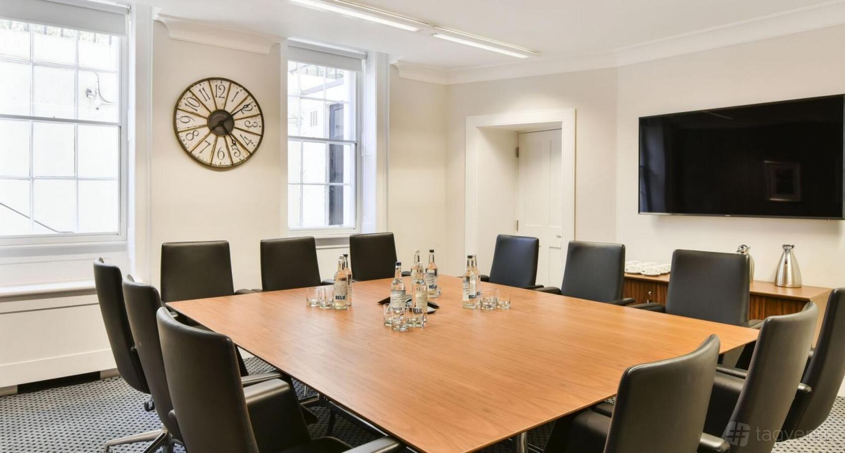 A meeting room with a square wooden table, black chairs, large wall clock, and mounted screen at 21 Gloucester Place.