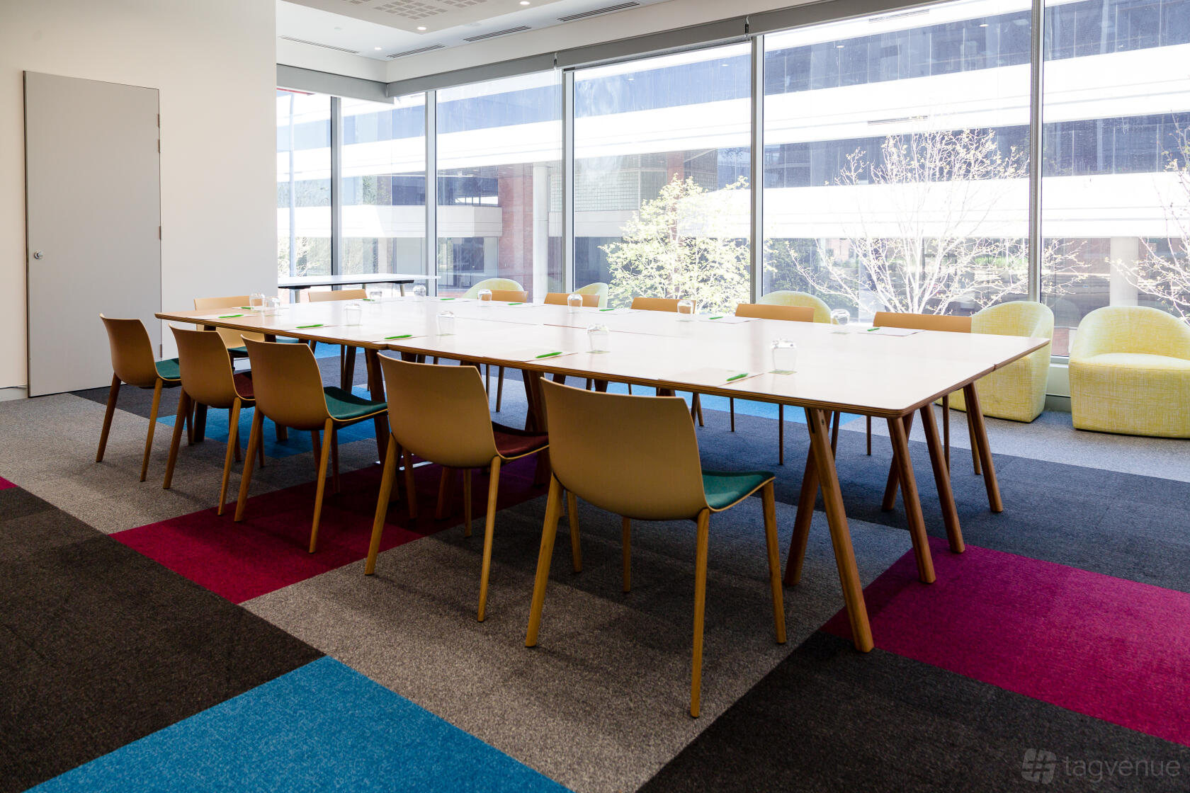 A hotel function room with floor-to-ceiling windows, rectangular tables in a U-shape, and colorful carpet at ibis Styles East Perth.