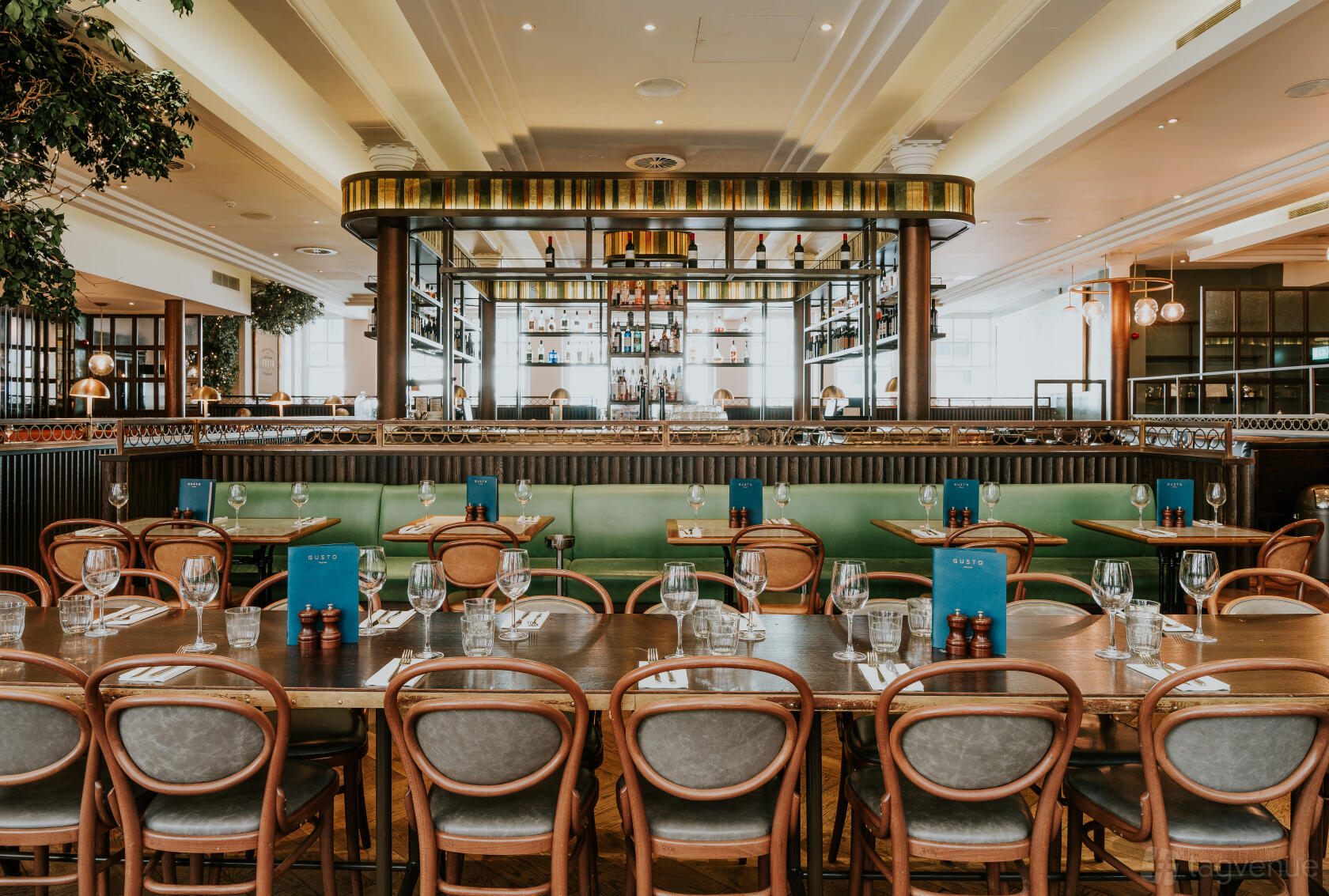 A restaurant with wooden tables set with glassware, green banquette seating, and a mirrored bar at Gusto Italian Manchester.