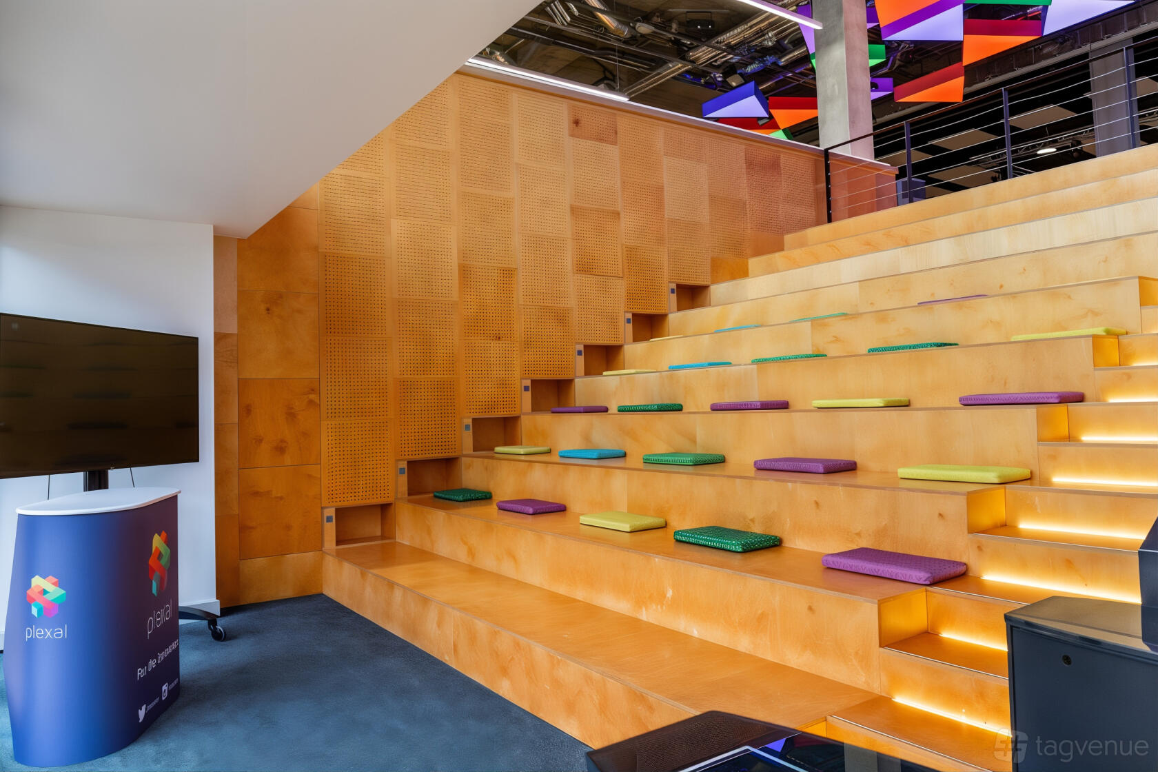 A training room with tiered wooden bleacher seating, colorful cushions, and a large screen at PLEXAL.
