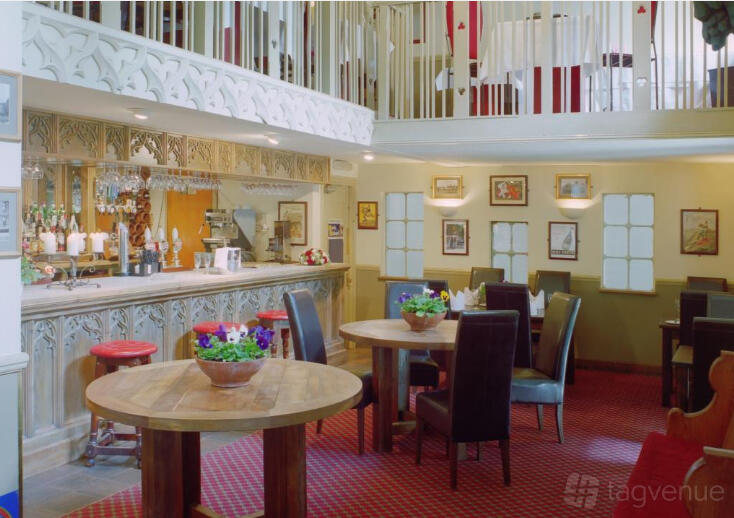 A bar in a banqueting hall with ornate railings, framed wall art, and round wooden tables at Le Gothique.