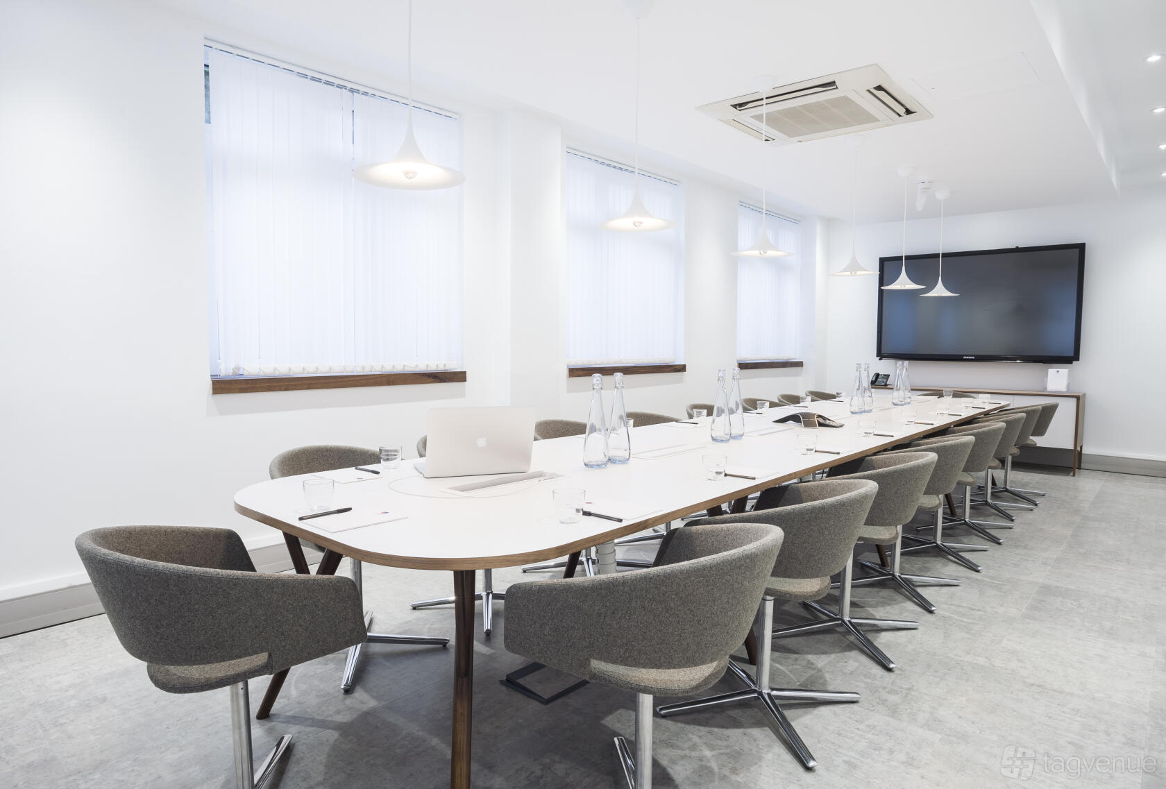 A meeting room with a long white conference table, modern gray chairs, pendant lights, and a wall-mounted screen at The Derby.