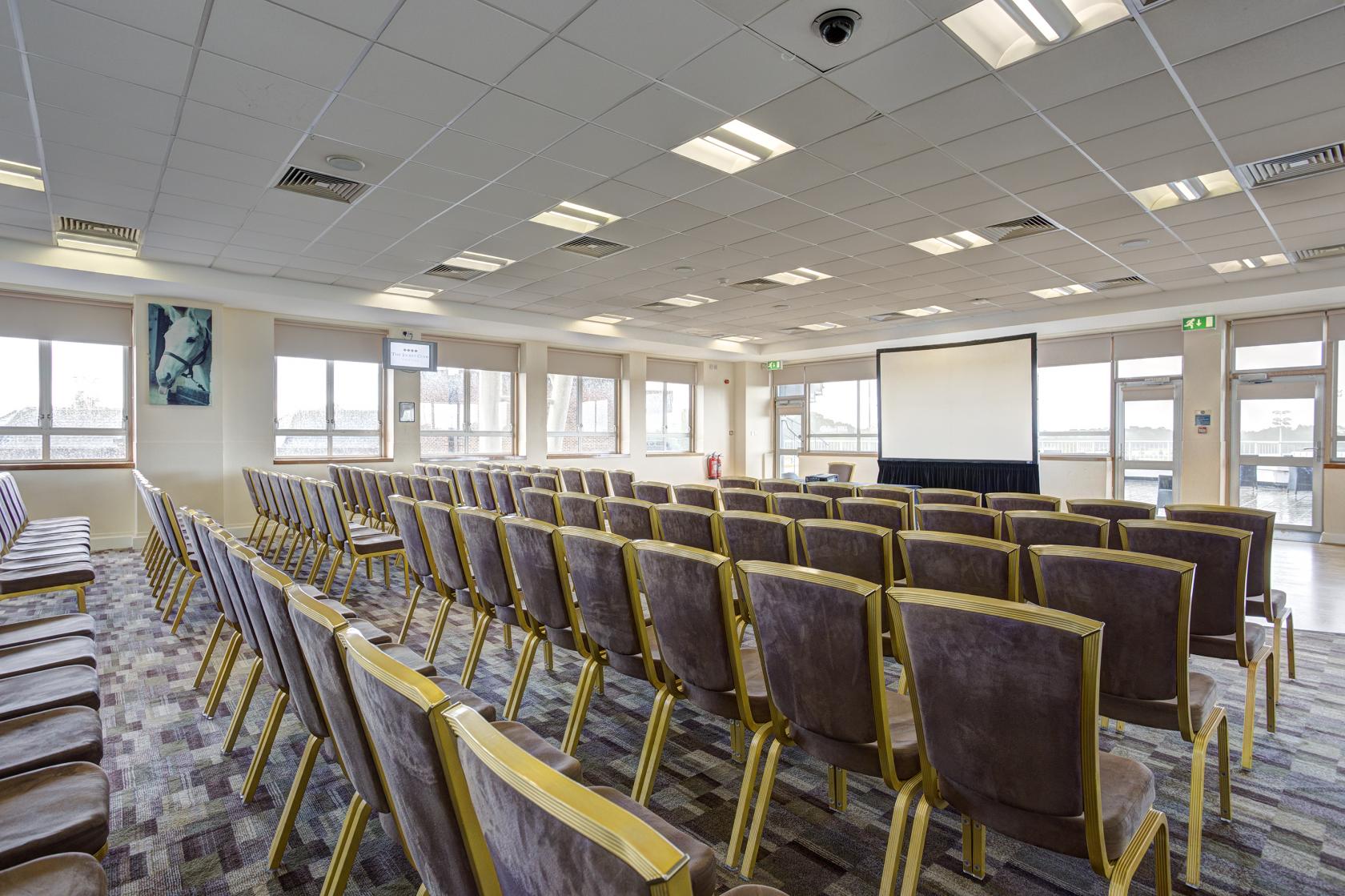 A function room with rows of cushioned chairs, projector screen, and large windows at Kempton Park Racecourse.