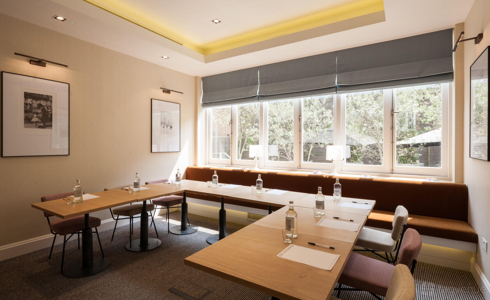 A hotel meeting room with large windows, natural light, wooden tables arranged in a U-shape, and cushioned bench seating at The Rockwell Hotel.