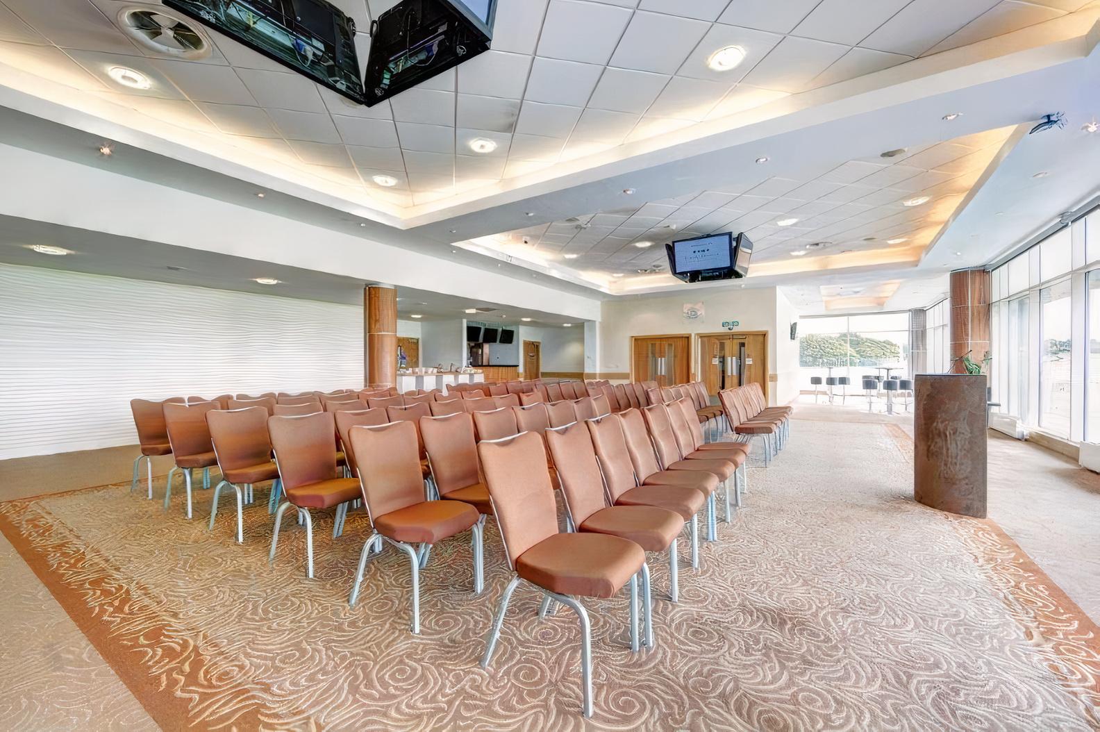 A meeting room with rows of upholstered chairs, patterned carpet, and large windows at Epsom Downs Racecourse.