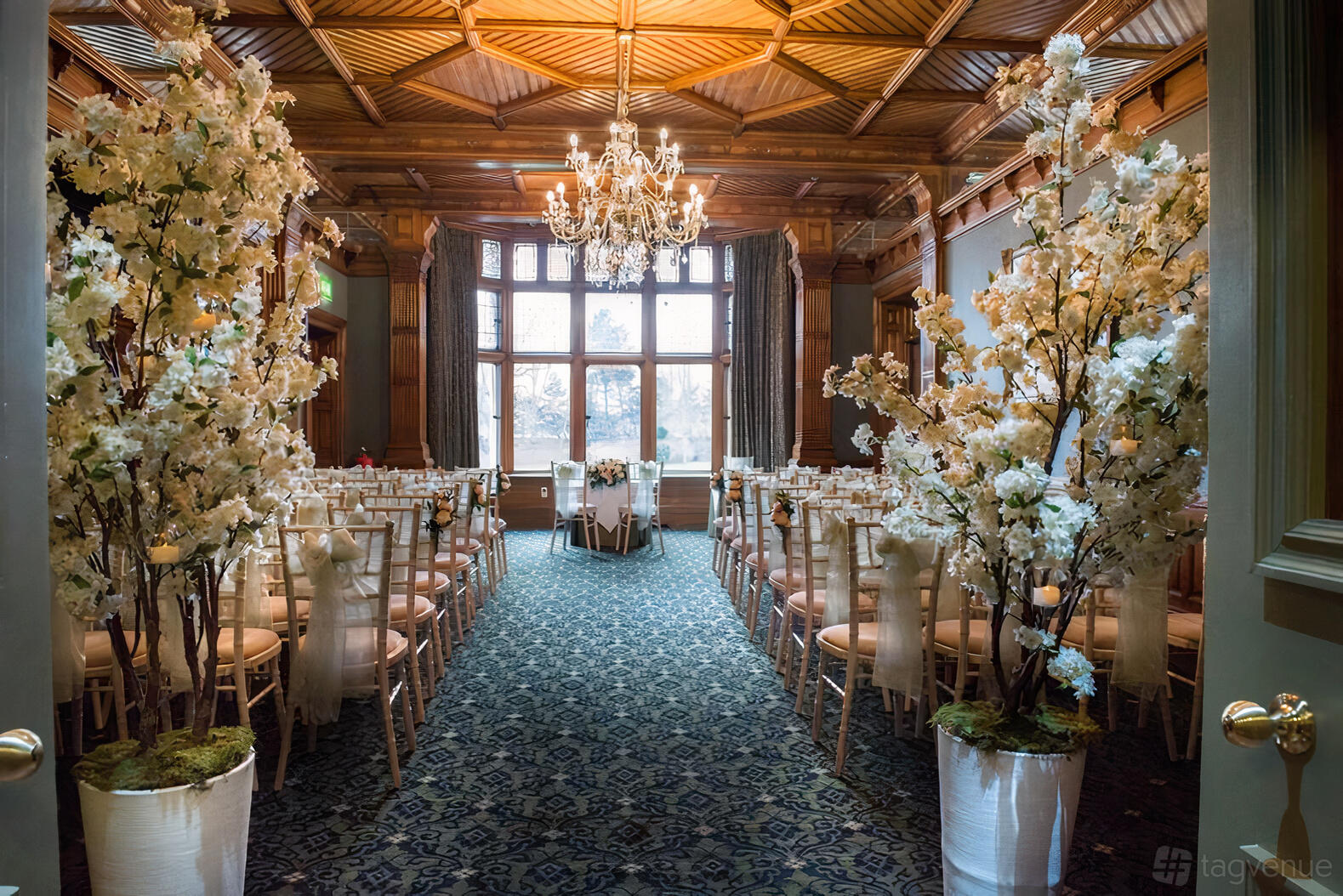 An event space with chandeliers, wooden ceiling, large windows, and aisle lined with white floral arrangements at Woodlands Park Hotel.