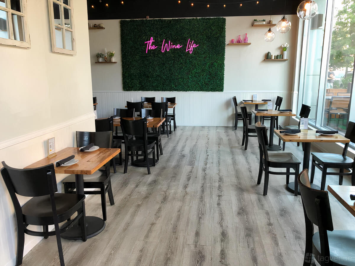 A bar with wooden tables, black chairs, large windows, and a neon sign on a green wall at Verre Wine Bar.