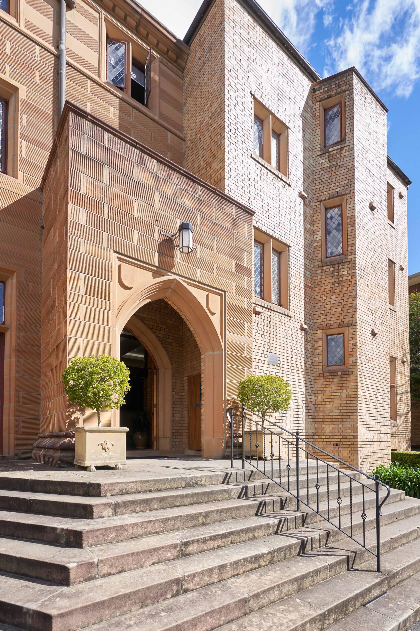 An academic building with arched sandstone entrance, detailed brickwork, and manicured bushes at Sancta Sophia College.