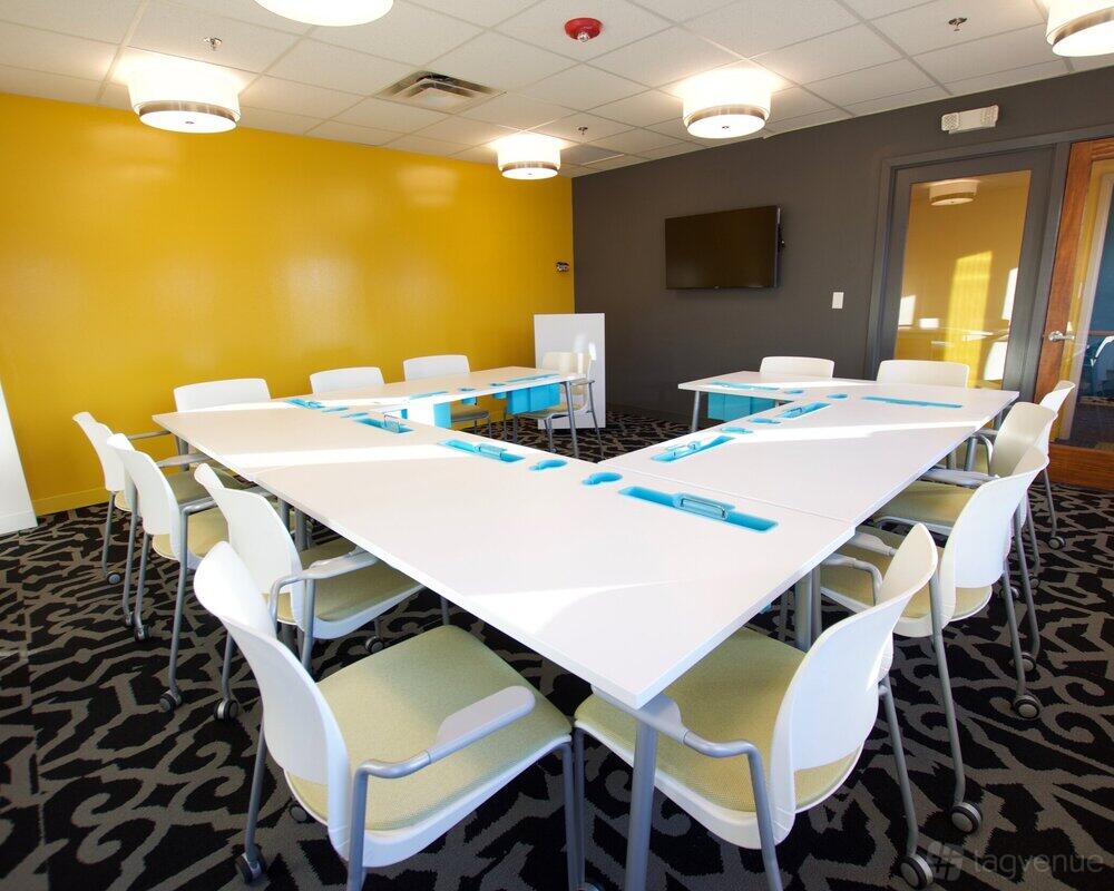 A meeting room with a U-shaped table arrangement, yellow accent wall, and mounted TV at 25N Coworking Geneva.