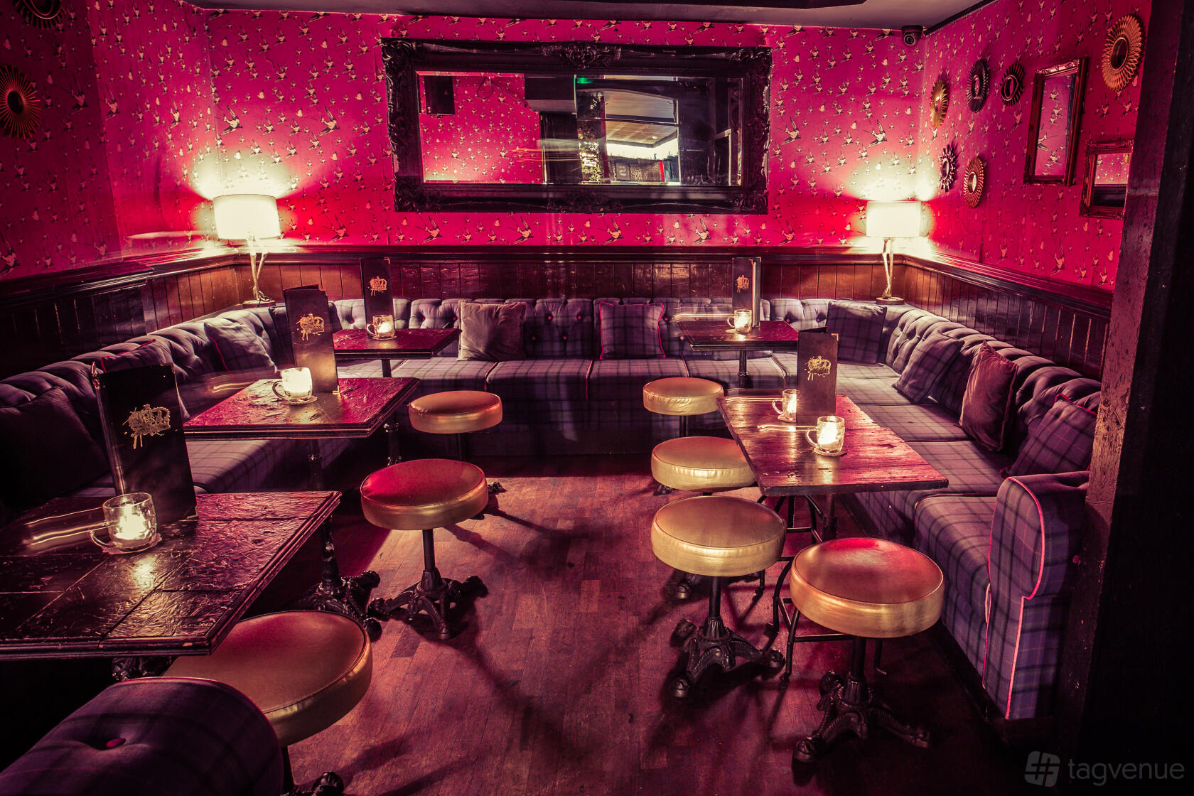 A cocktail bar with plush plaid banquettes, round stools, candlelit tables, and red patterned wallpaper at The Cocktail Club Goodge Street.