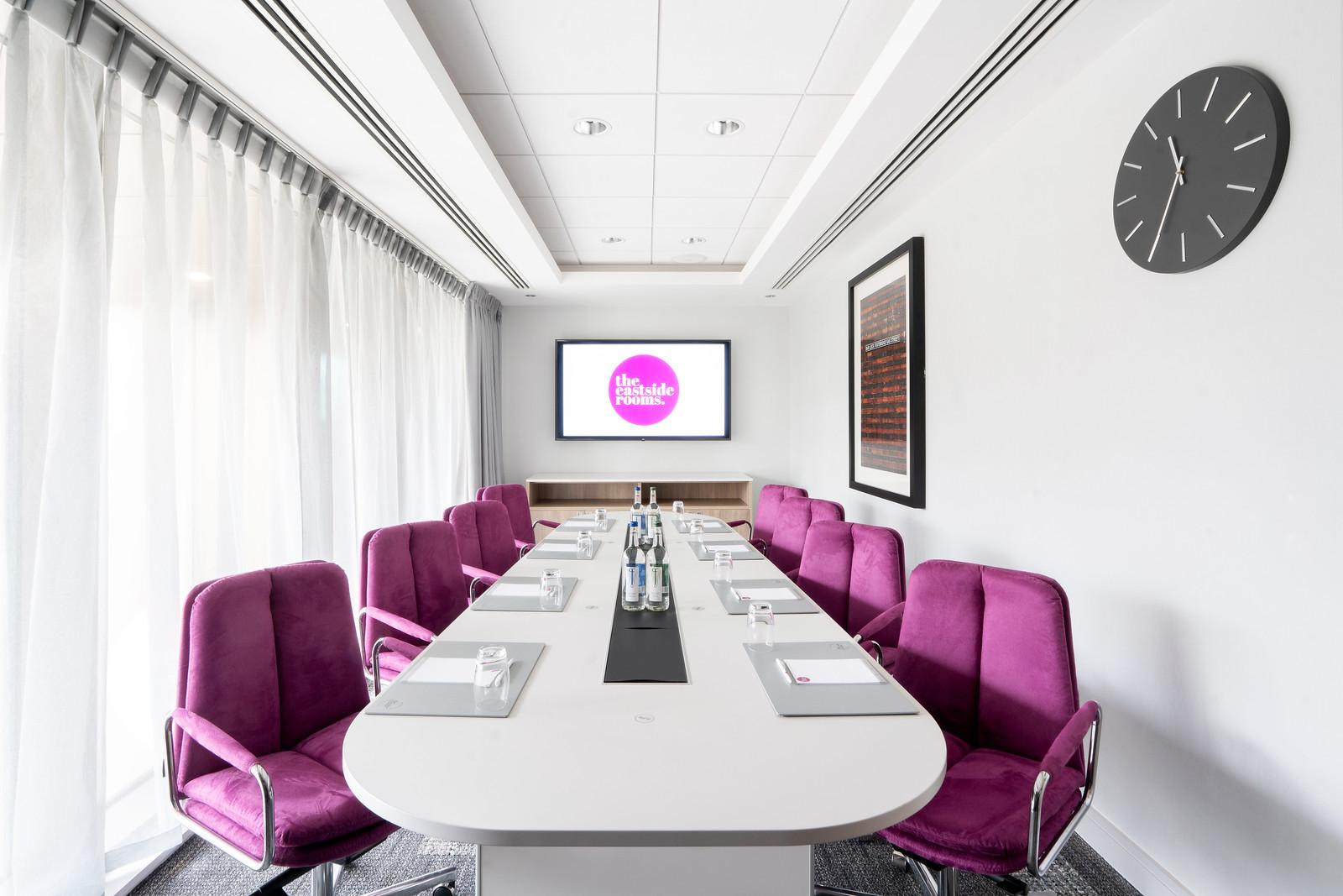 A boardroom with magenta chairs, a white oval table, and a wall-mounted display screen at The Eastside Rooms.