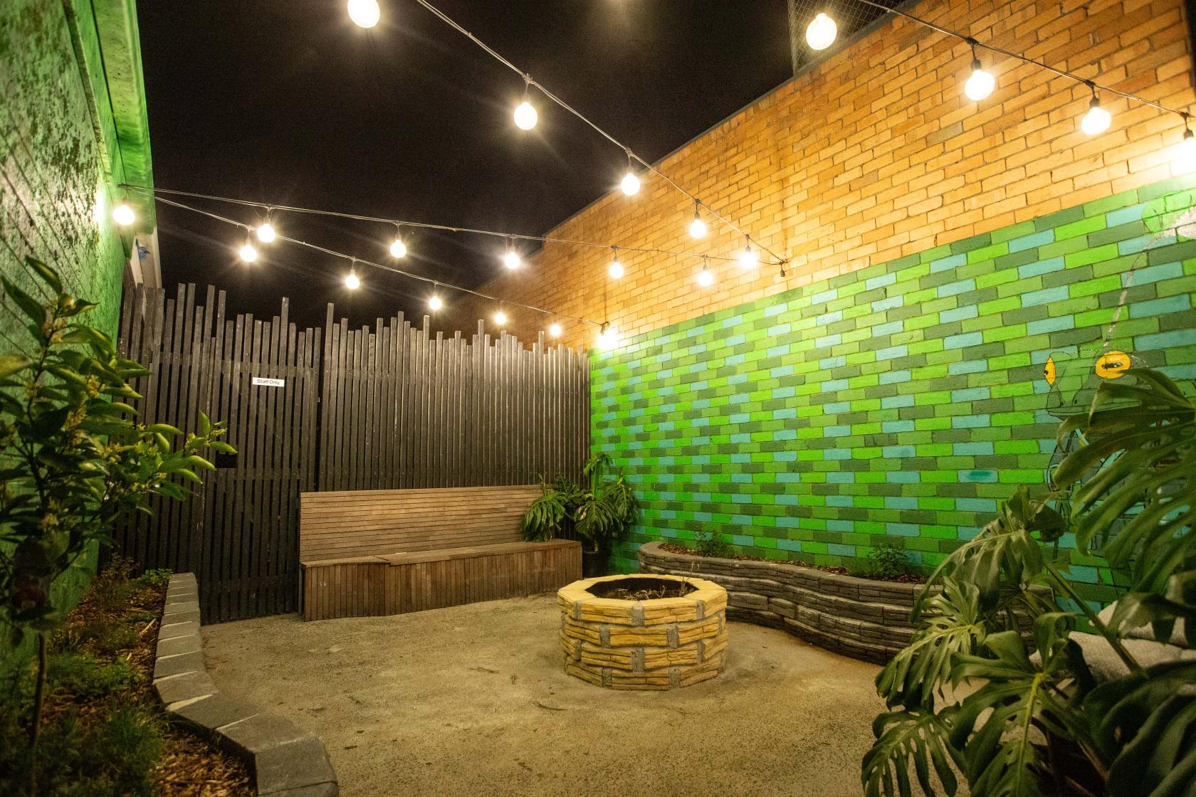 An outdoor bar courtyard with string lights, a brick fire pit, and colorful green brick walls at Lucky Schmuck.
