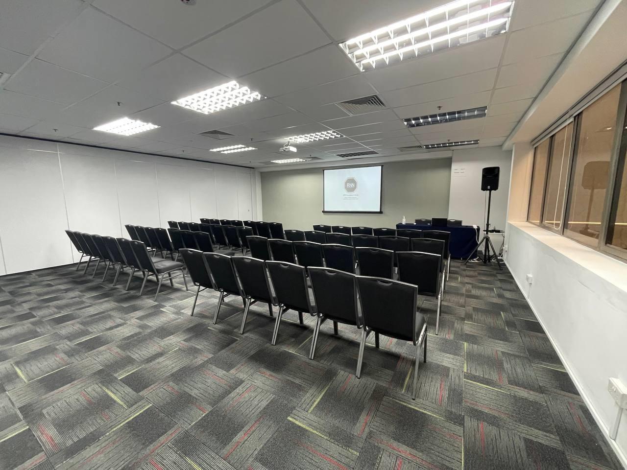 An auditorium with rows of black cushioned chairs, a projector screen, and fluorescent lighting at RNN Conference Centre.