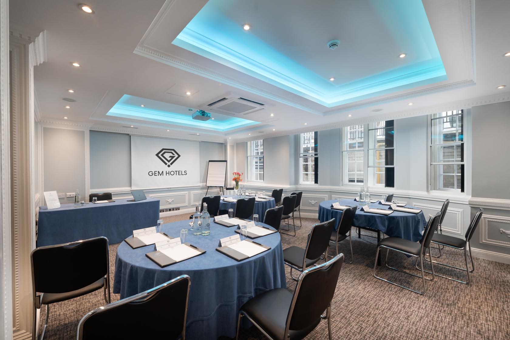 An event space with round tables covered in blue linens, modern ceiling lighting, and large windows at Fitzrovia Hotel.