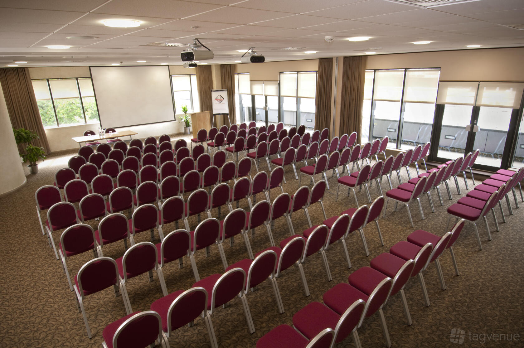 An event space with rows of red cushioned chairs facing a projector screen and windows at Future Inns Hotel Bristol.