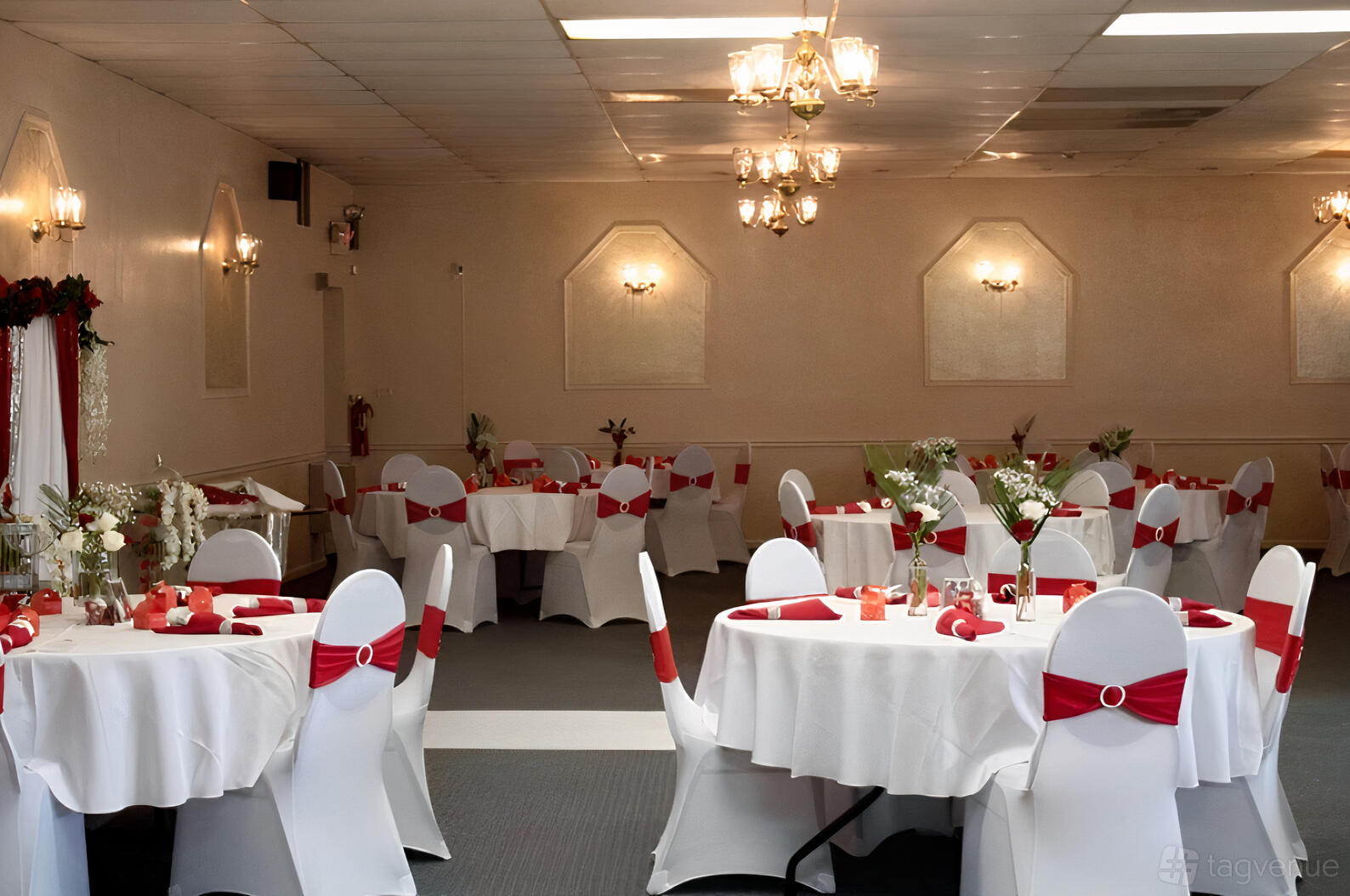 A banquet hall with round tables draped in white linens and red chair sashes under chandelier lighting at Wedding Kiss Ballroom.