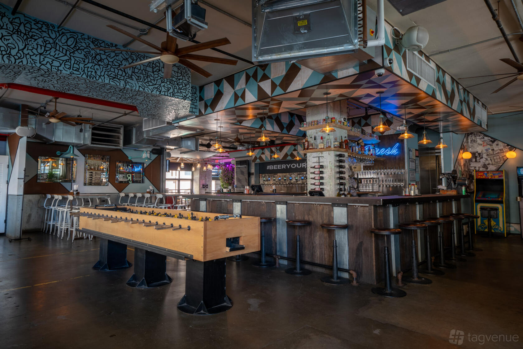 A pub with a central bar, foosball table, industrial lighting, and geometric ceiling decor at Randolph Beer DUMBO.