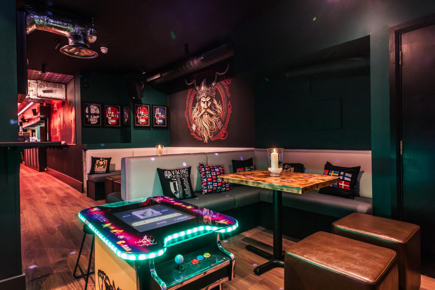 A bar with Viking-themed wall art, a retro arcade table, and booth seating at Nordic Bar.