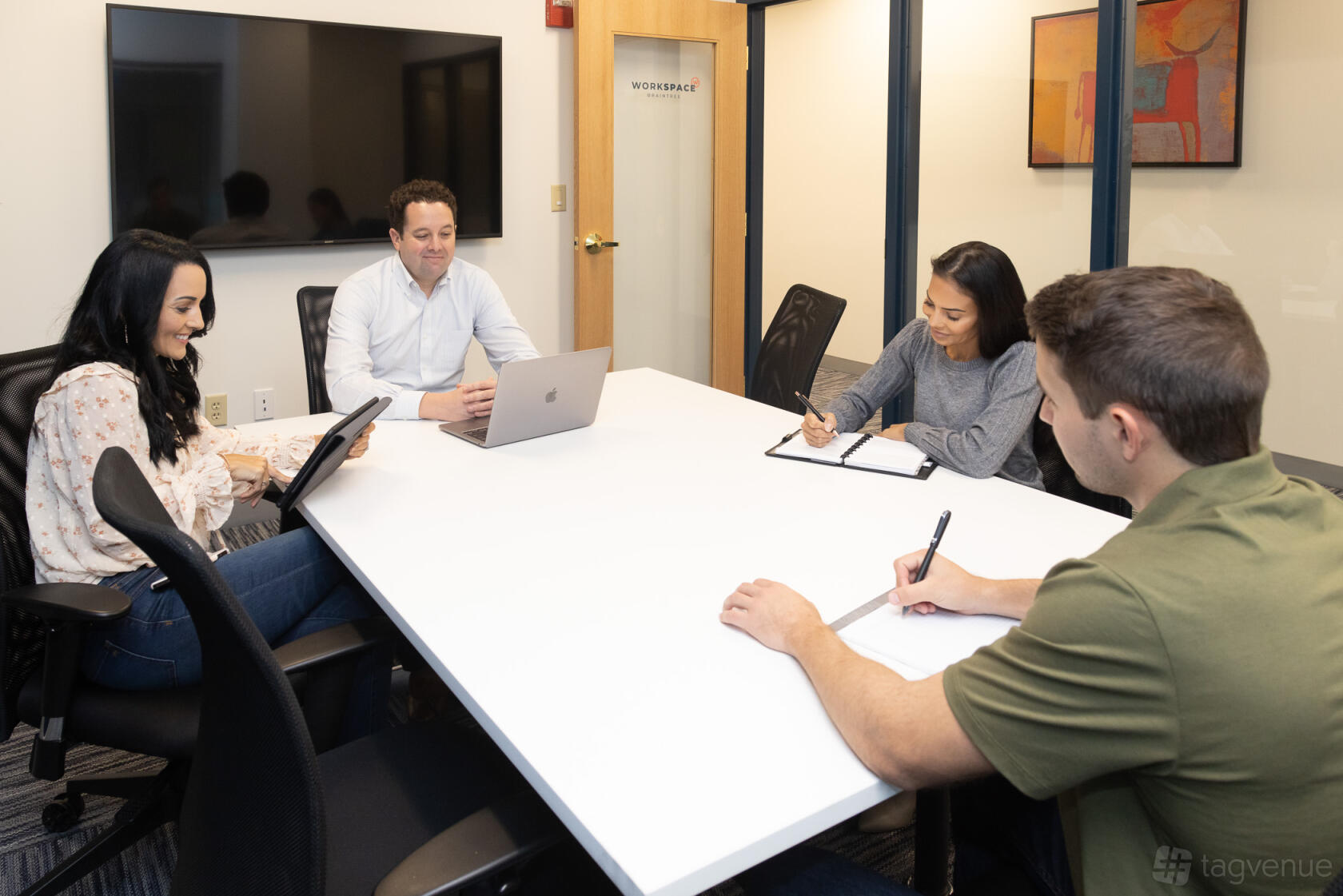 A meeting room with a large white conference table, black office chairs, and a wall-mounted screen at Workspace Braintree.