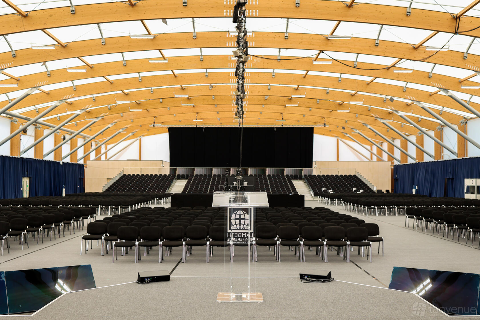 An event venue with rows of black chairs, a central stage, and a curved wooden beam ceiling at University of Kent.