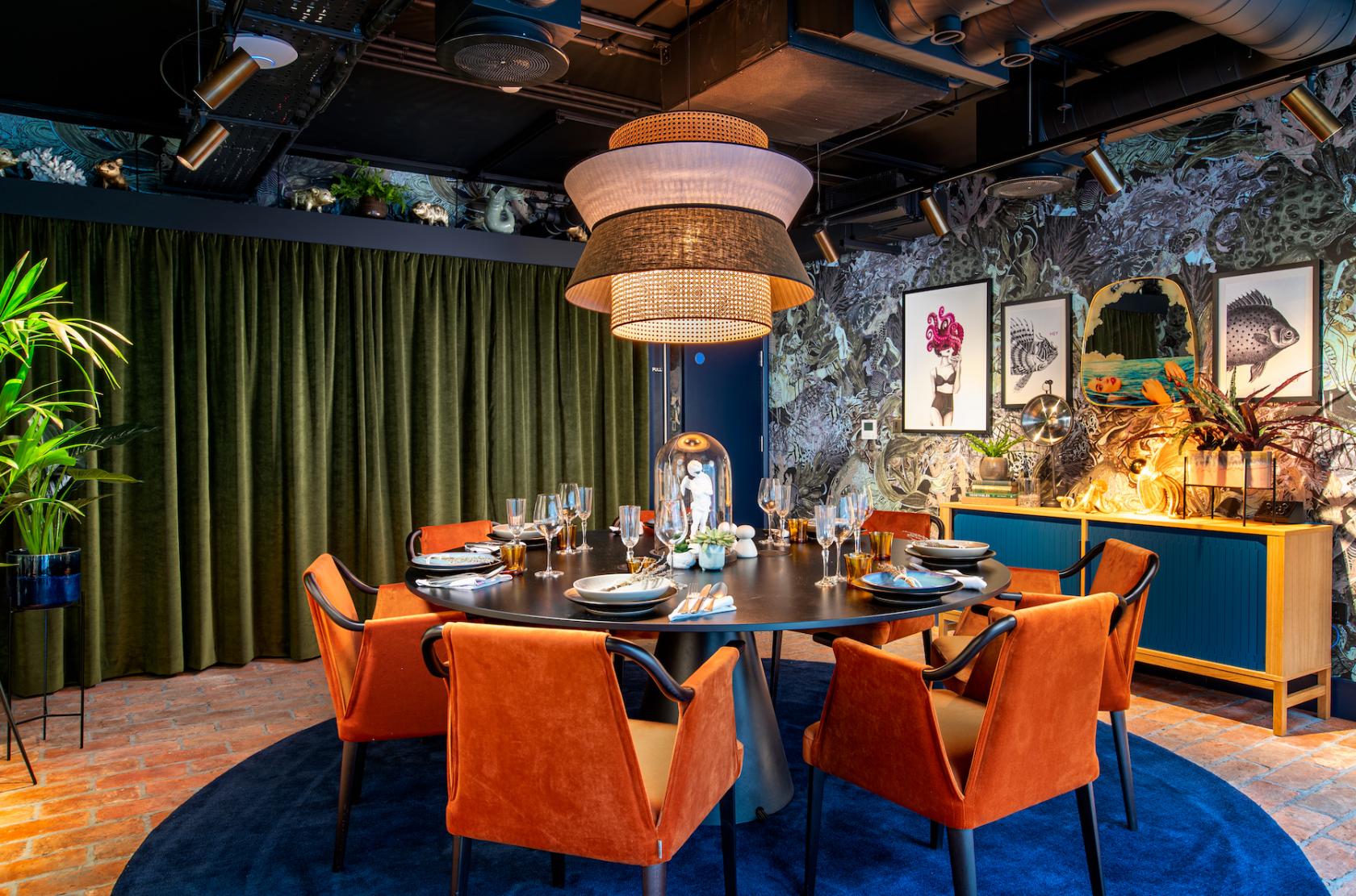 A dining room with an oval wooden table, orange velvet chairs, and a statement pendant light at Yotel Manchester Deansgate.