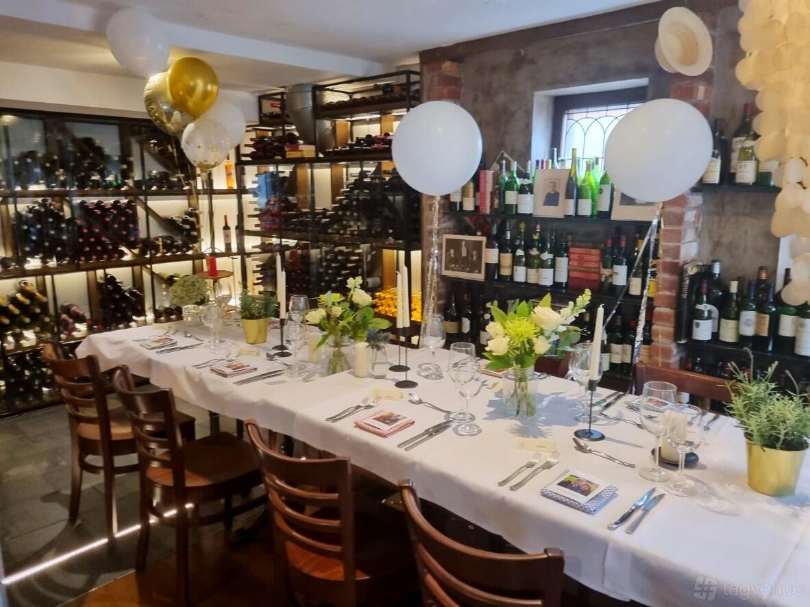 A dining room with a long table set with white linens, floral centerpieces, balloons, and a wine rack wall at Gazette Battersea.