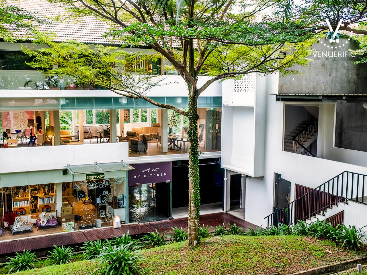 A restaurant with glass walls, tree-shaded outdoor walkway, and modern facade at EAT AT MY KITCHEN.