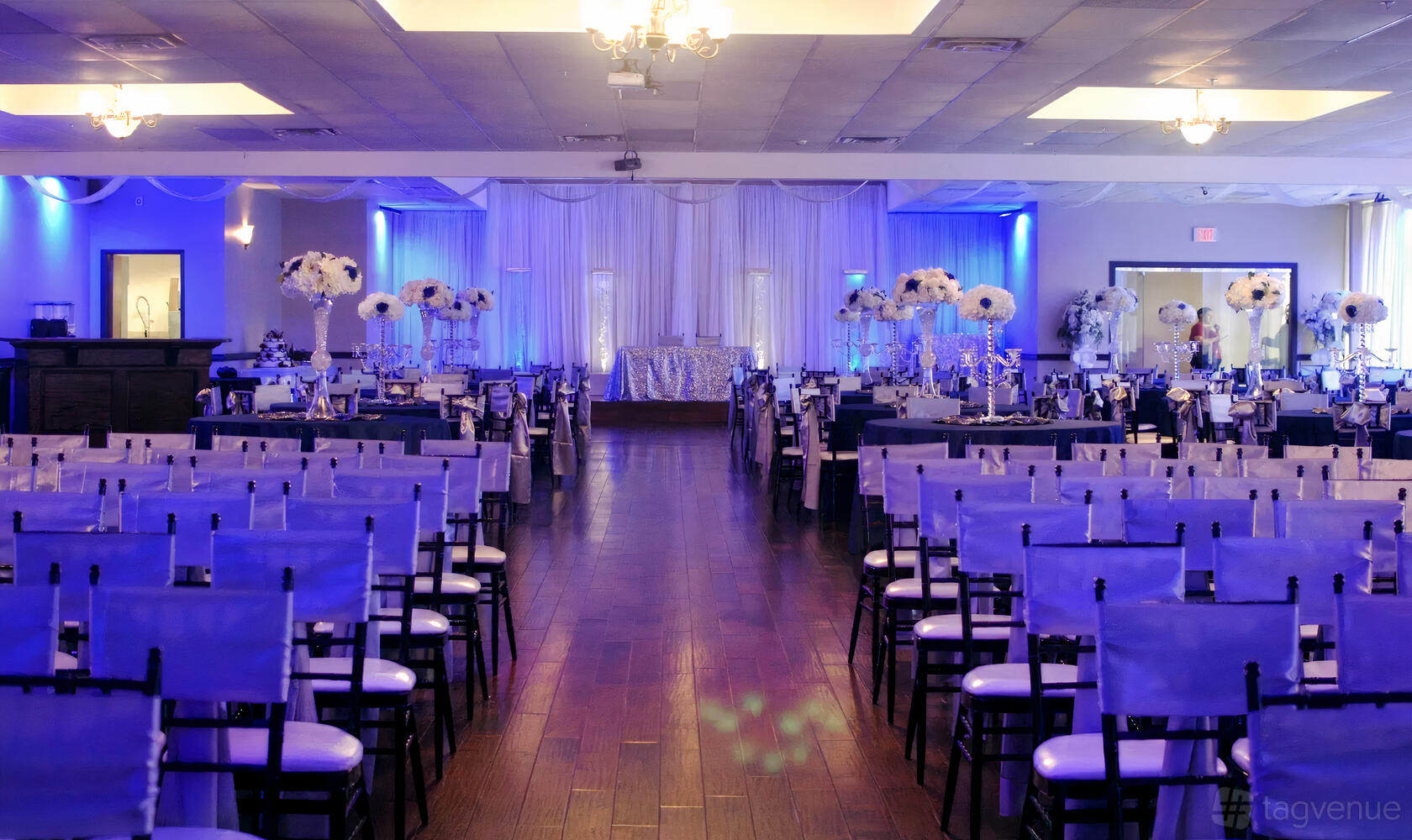 A hall with rows of chairs, wood flooring, blue accent lighting, and floral centerpieces at The Oasis Ballroom.