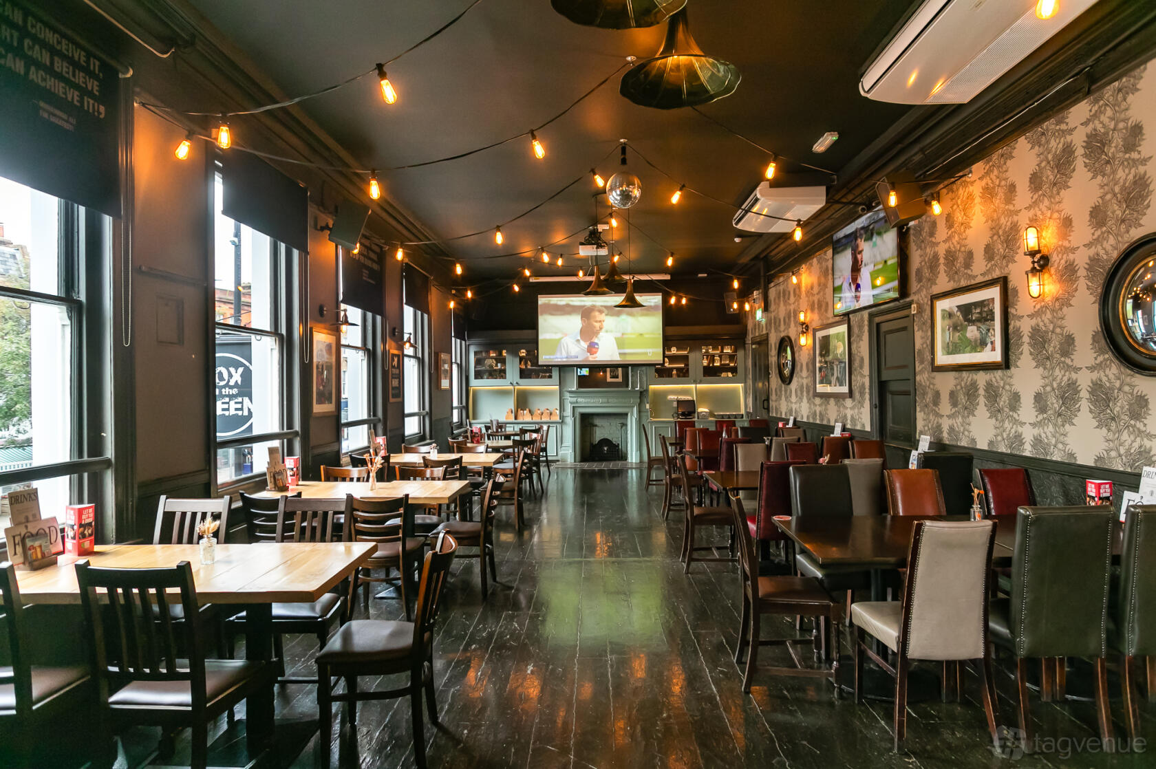 A pub bar with dark wood floors, patterned wallpaper, string lights, and a projector screen at Fox On The Green.