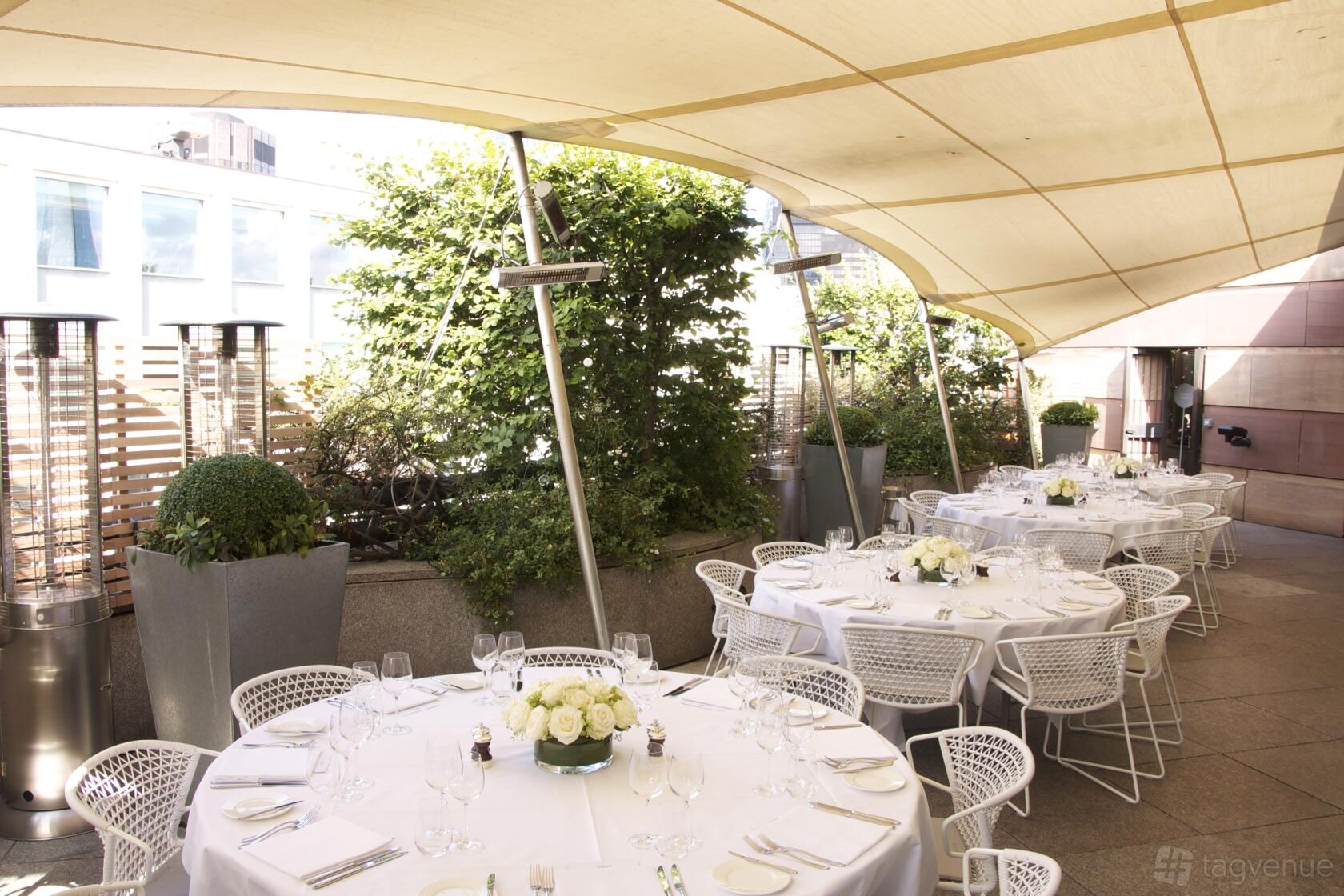 A restaurant terrace with round tables dressed in white linens, white chairs, planters, and canopy cover at Coq D'Argent.