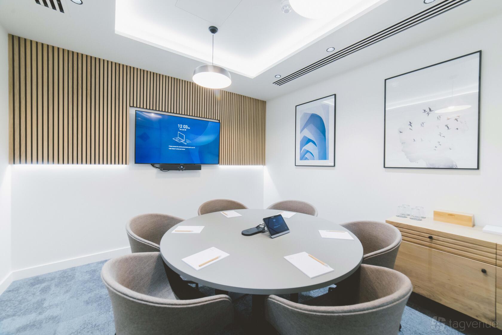A boardroom with a round table, six grey chairs, wall-mounted screen, and wood panel accents at Orega Marlow.