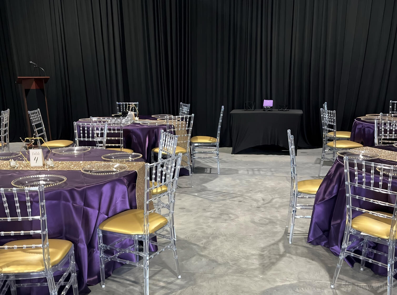 An event venue with round tables draped in purple linens, clear chairs, and a podium at SW Party and Events.