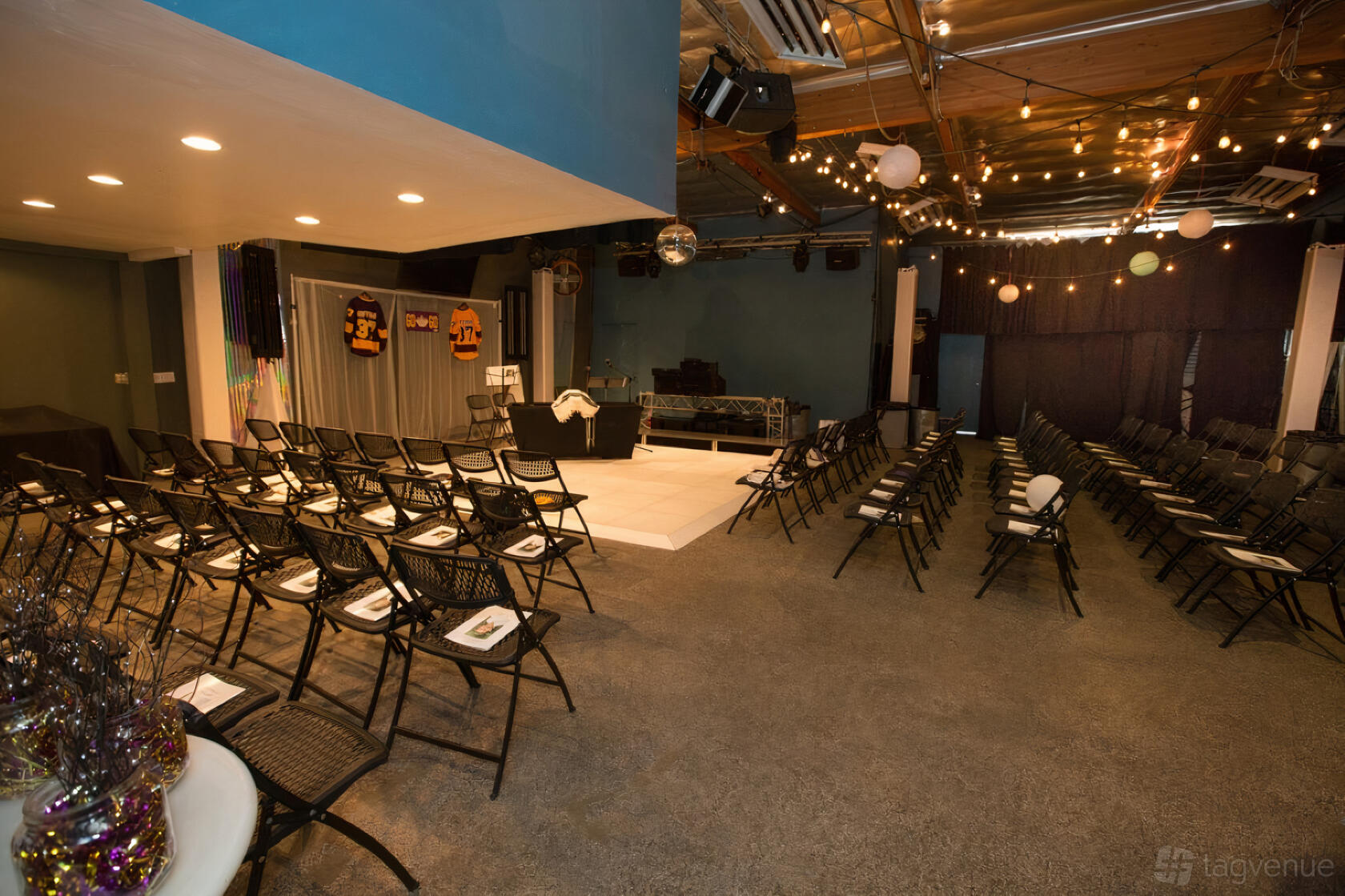 An event venue with rows of black folding chairs, a small stage, and string lights at Club Energy.