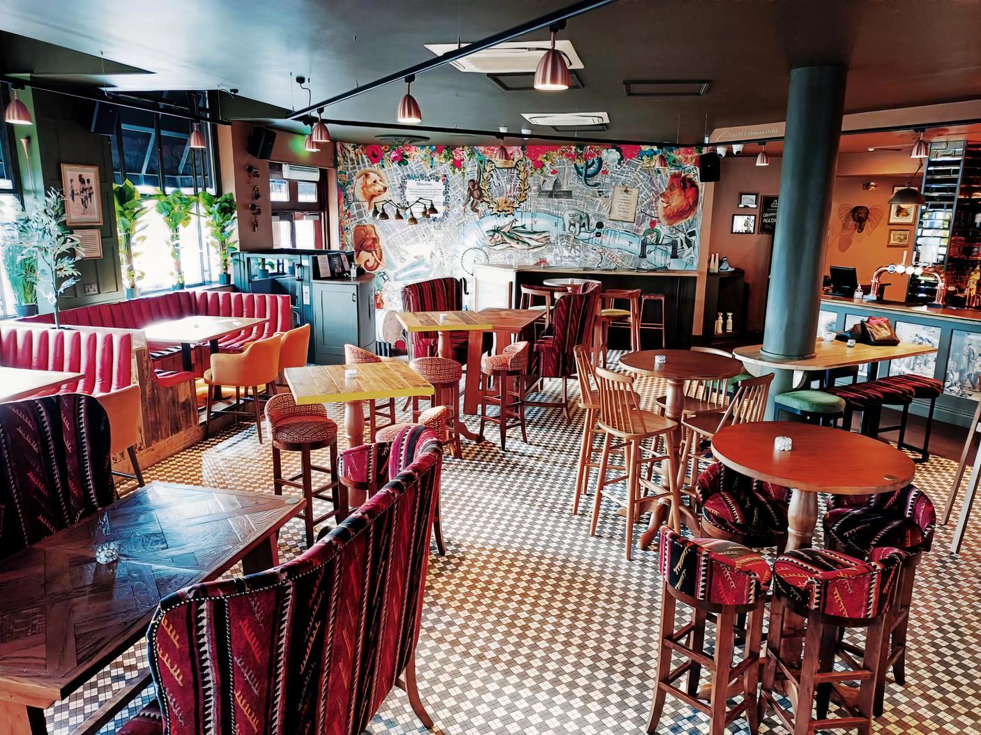 An underground pub space with patterned tile floors, upholstered chairs, and mural-covered walls at Traitors Gate Tower Hill.