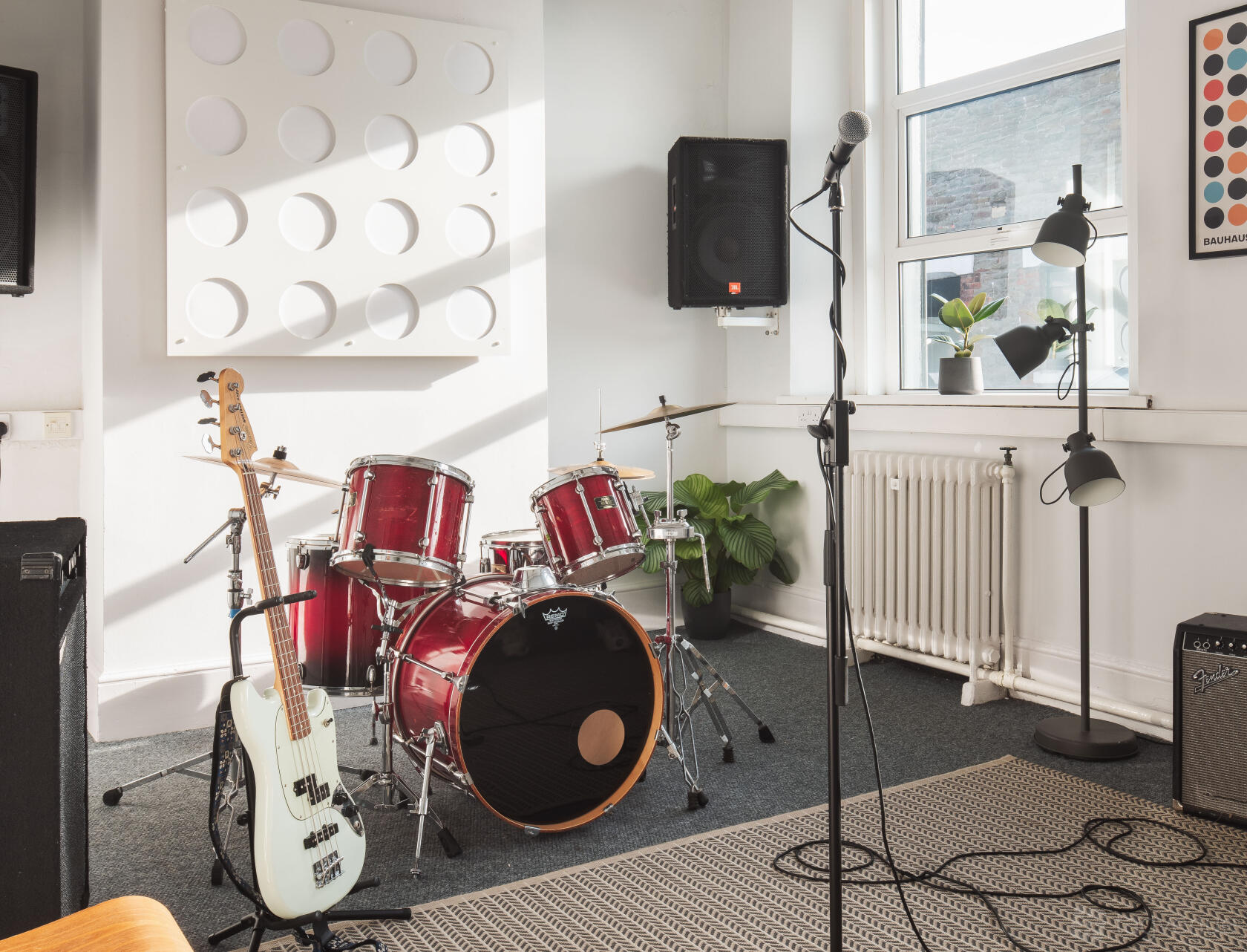 A rehearsal studio with a red drum kit, microphone stand, and guitars near sunlit windows at Factory Studios.