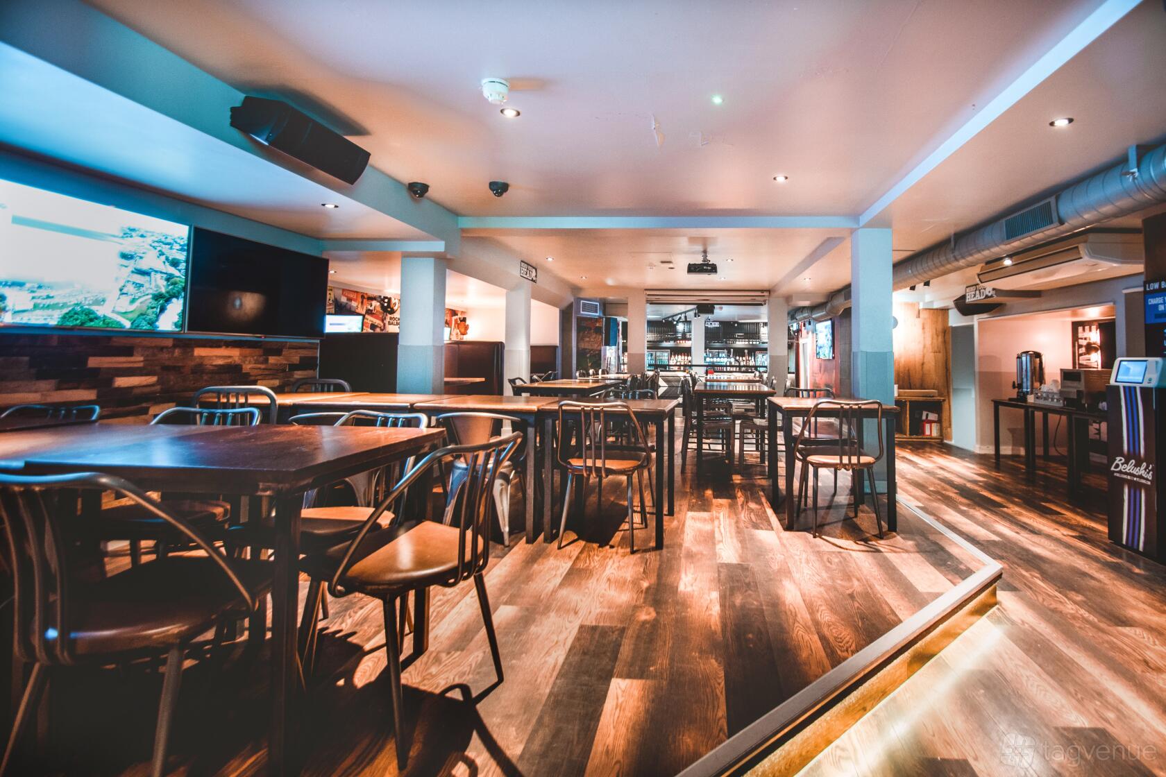 An event space with wooden floors, black chairs, and multiple screens at Belushi's & The Dugout London Bridge.