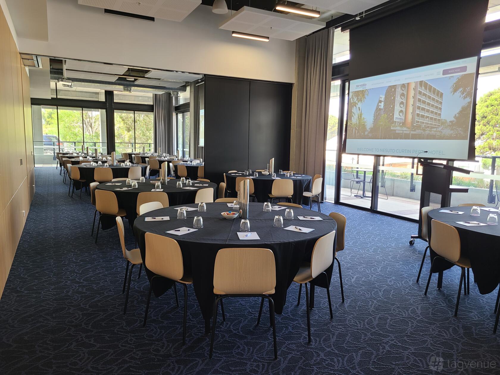 A function room with round tables covered in black linens, floor-to-ceiling windows, and a projector screen at Nesuto Curtin Perth Hotel.