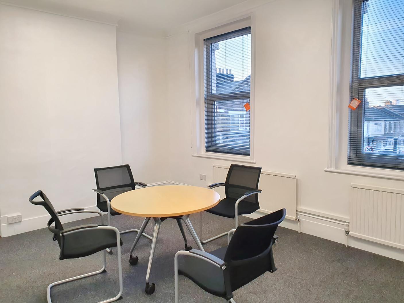 A coworking space with four black mesh chairs around a round wooden table by large windows at SBN West Ham Lane.