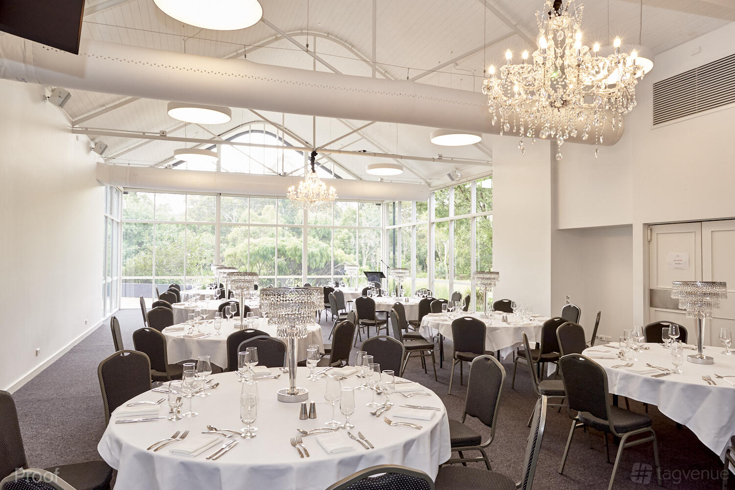 A hall with round tables draped in white linens, crystal chandeliers, and floor-to-ceiling windows at No 1 Events- Malvern.