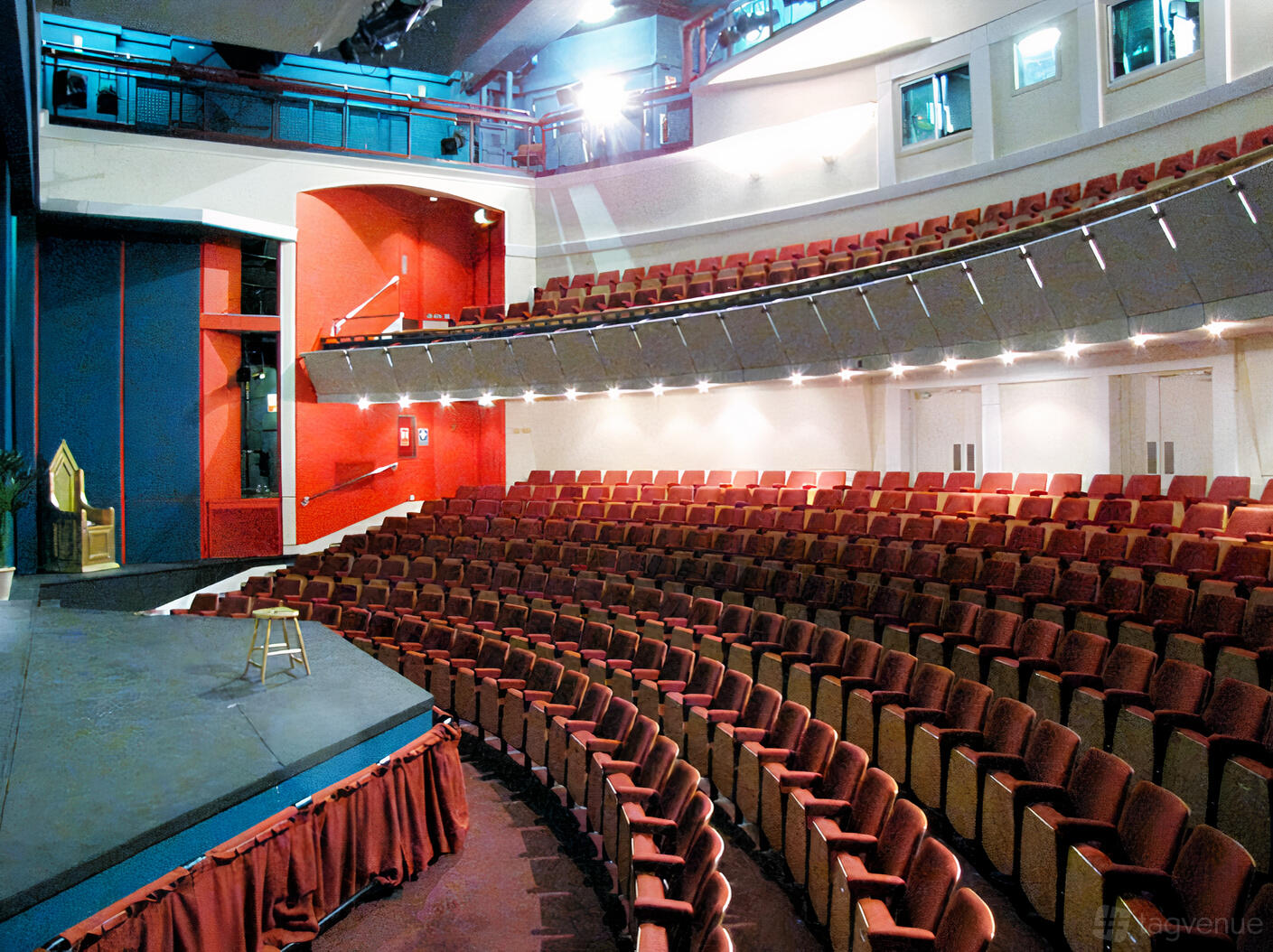 A school theatre with tiered red seating, curved balcony, and stage with a stool at Harrow School’s Ryan Theatre.