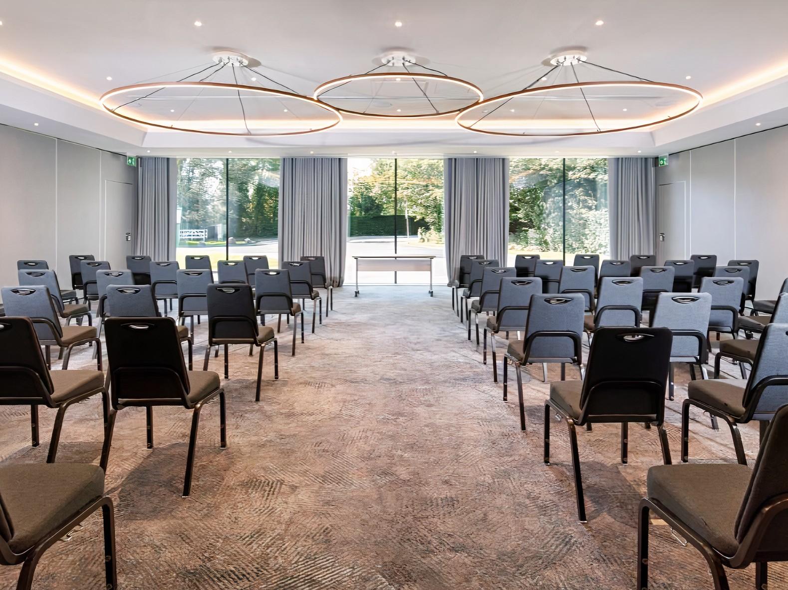 A hotel meeting room with rows of black chairs, large windows, and circular ceiling lights at Holiday Inn Shepperton.