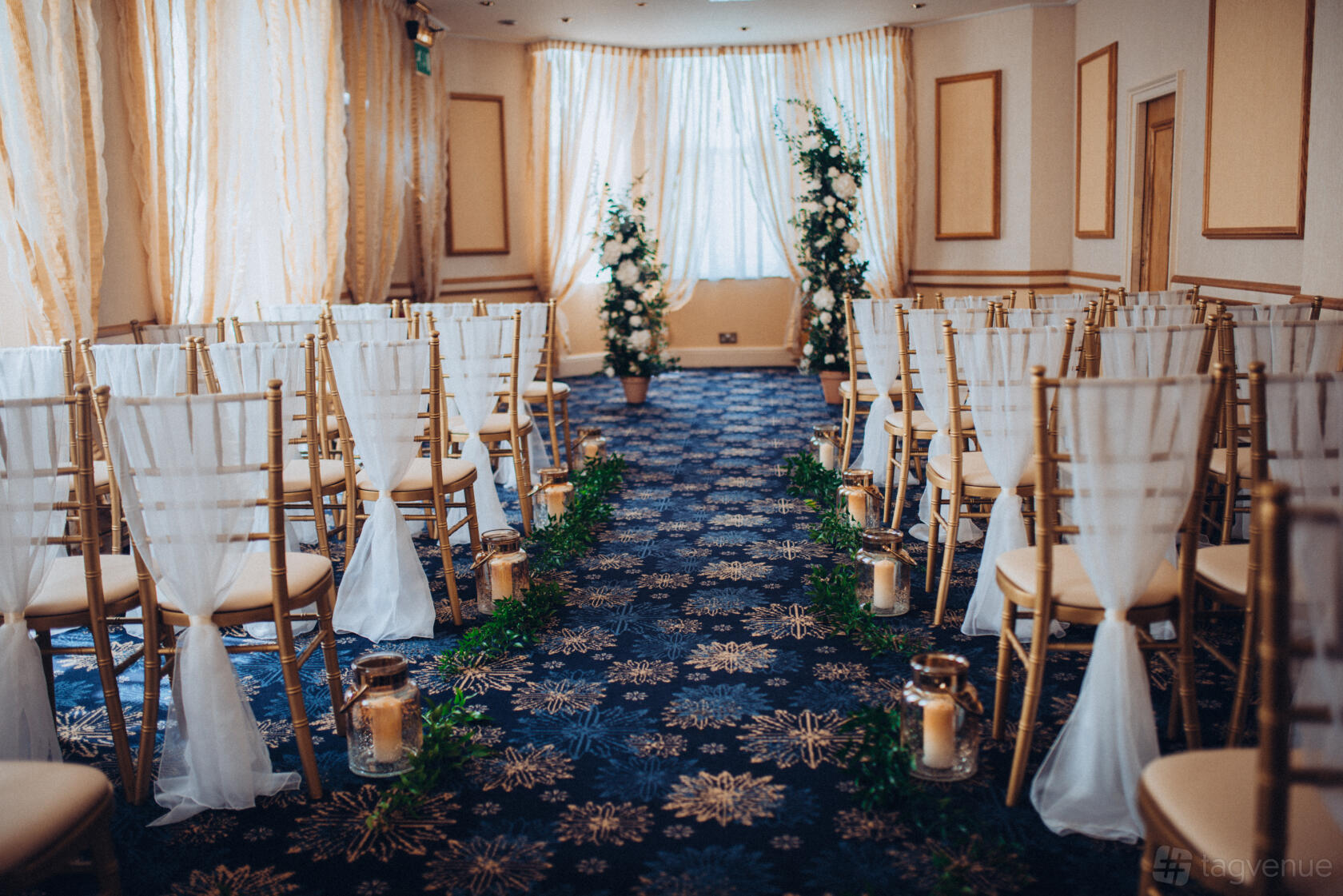 An event space with rows of gold chairs, white fabric draping, and floral aisle decor at New Continental Hotel.