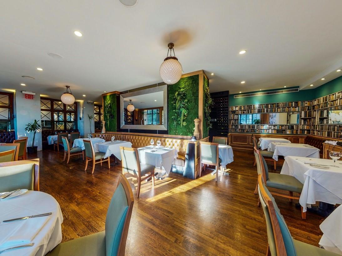 A dining room with wood floors, white tablecloths, green accent wall, and large mirror at Tudor City Steakhouse.