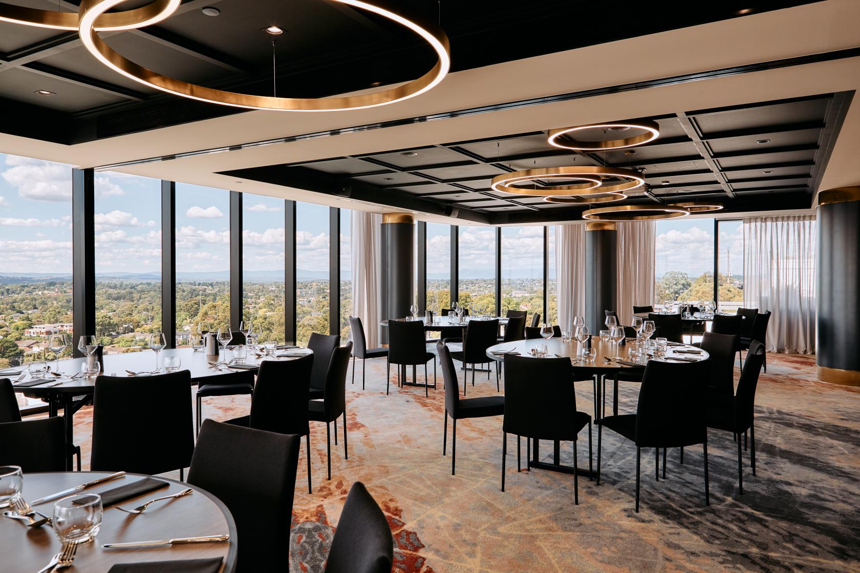 An event space with floor-to-ceiling windows, circular pendant lights, and round tables at Mercure Melbourne Doncaster.