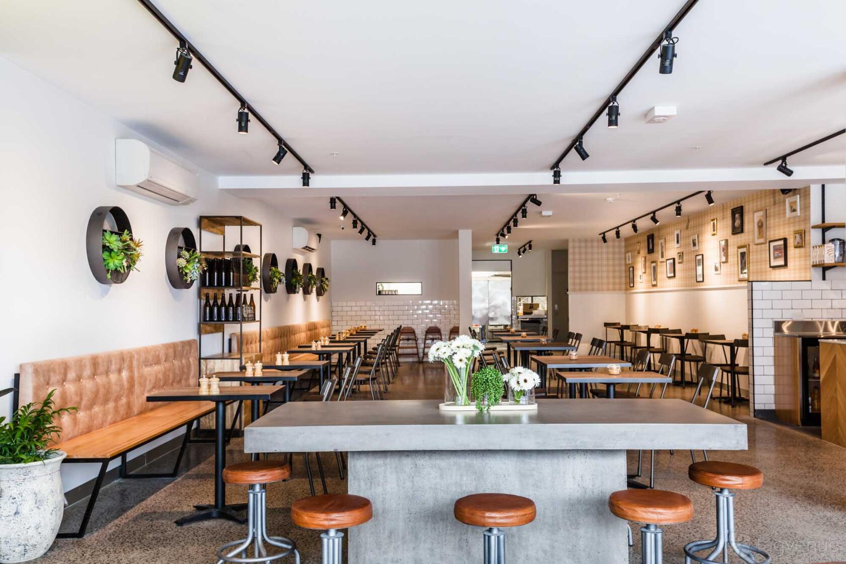 A cafe with long wooden tables, leather banquettes, framed wall art, and indoor plants at Sycamore Meadows Melbourne.