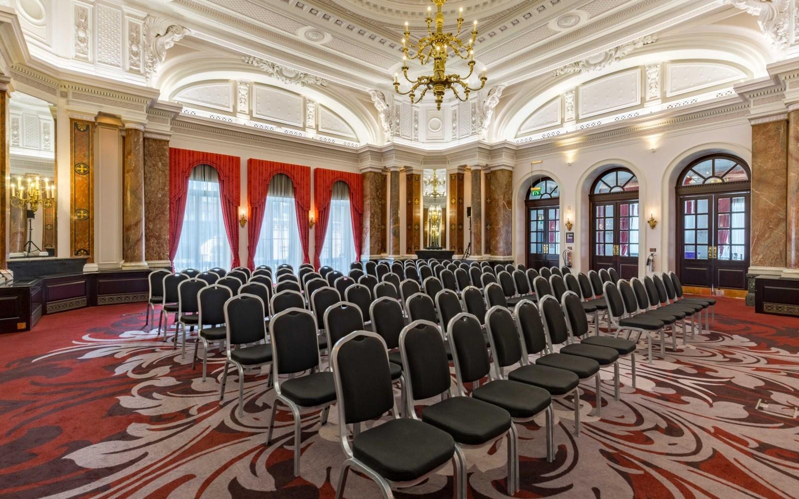 A hotel ballroom with arched windows, red curtains, ornate ceiling moldings, and rows of chairs at Clermont Charing Cross.