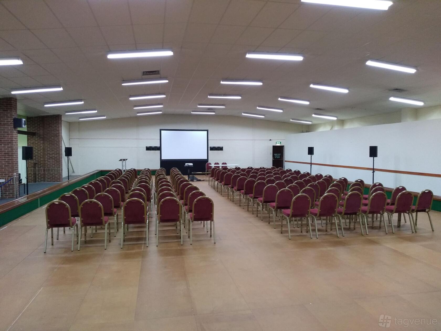 An indoor hall with rows of red cushioned chairs facing a large projection screen at Cambridge Chesterton Indoor Bowls Club.