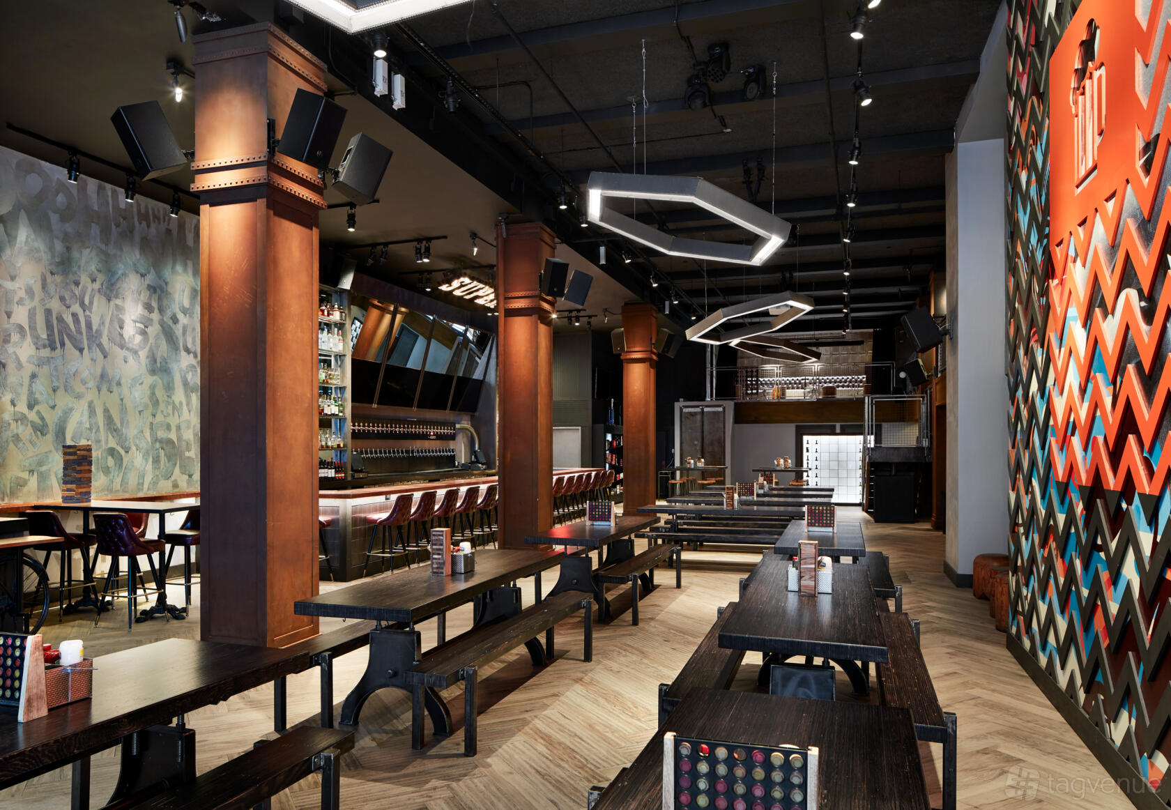 A bar with long communal wooden tables, geometric lighting, and a colorful mural at Clinton Hall 36th Street.